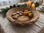 Shallow wicker bread basket with gingerbread stars, a heart cookie and tangerines on a linen tablecloth among candles and fir branches.