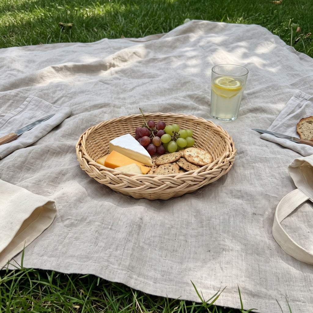 Round willow serving basket on a linen picnic blanket in the grass, filled with brie, cheddar, grapes and crackers next to a glass of lemonade.