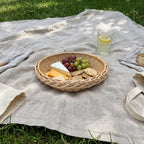 Round willow serving basket on a linen picnic blanket in the grass, filled with brie, cheddar, grapes and crackers next to a glass of lemonade.