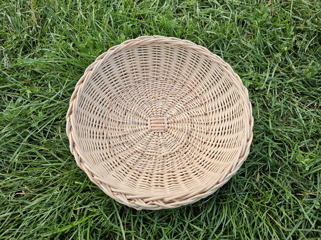 Round handwoven willow tray photographed from above on green grass, showing the even radial weave and neat braided rim.
