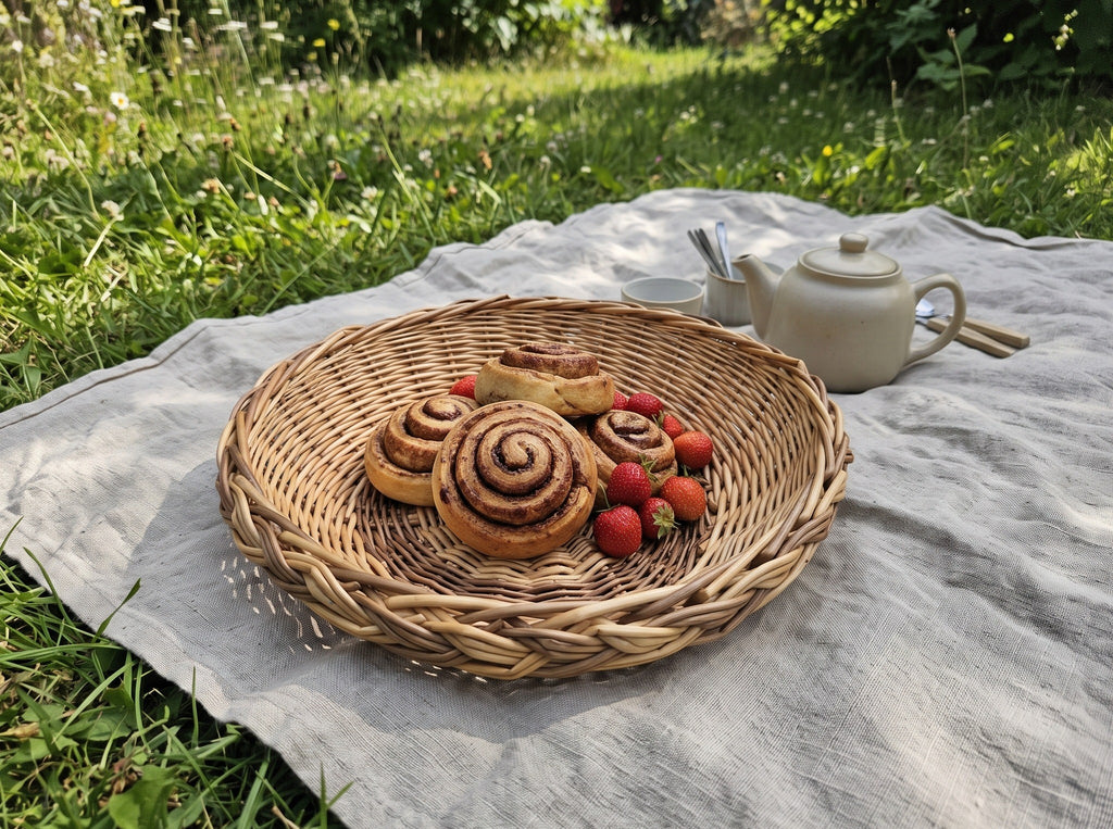 Handwoven wicker bread basket with cinnamon rolls and strawberries on a picnic blanket in the garden beside a ceramic teapot.