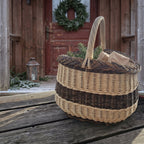 Willow market basket with twisted handle holding small wrapped gifts and fir sprigs on a wooden porch by a door with a Christmas wreath
