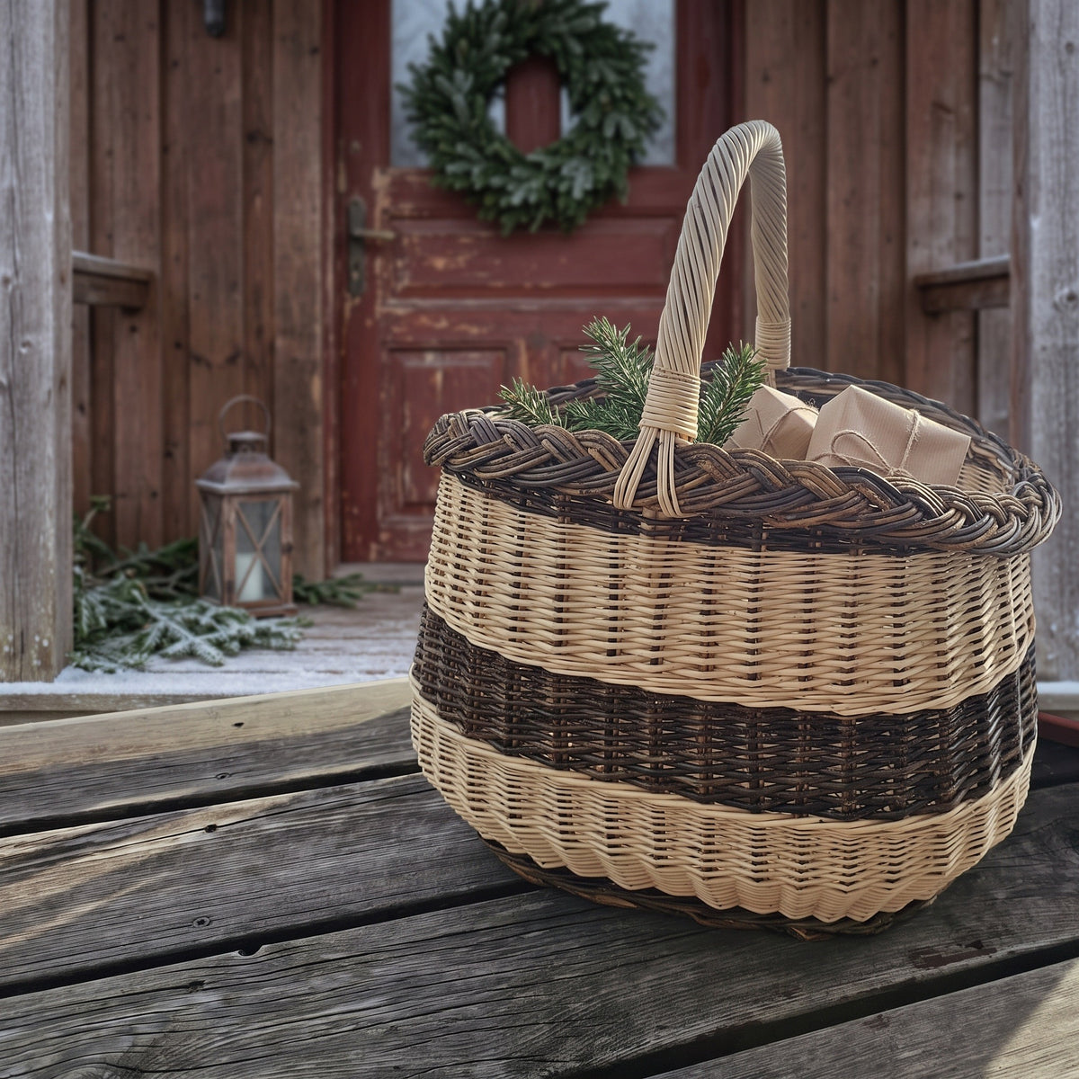 Willow market basket with twisted handle holding small wrapped gifts and fir sprigs on a wooden porch by a door with a Christmas wreath