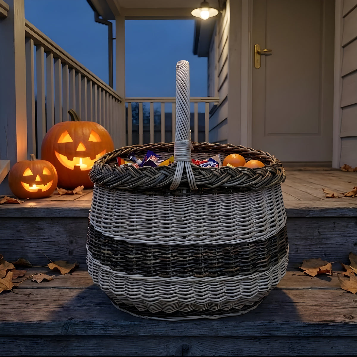 Oval wicker basket filled with sweets on a porch step between two lit jack-o-lanterns, ready for Halloween trick-or-treaters