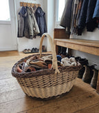 Large oval wicker storage basket with handle holding scarves and gloves on the floor of a hallway beside a coat rack