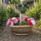 Large oval willow flower basket with a tall handle filled with fresh peonies and roses on a gravel path in a cottage garden