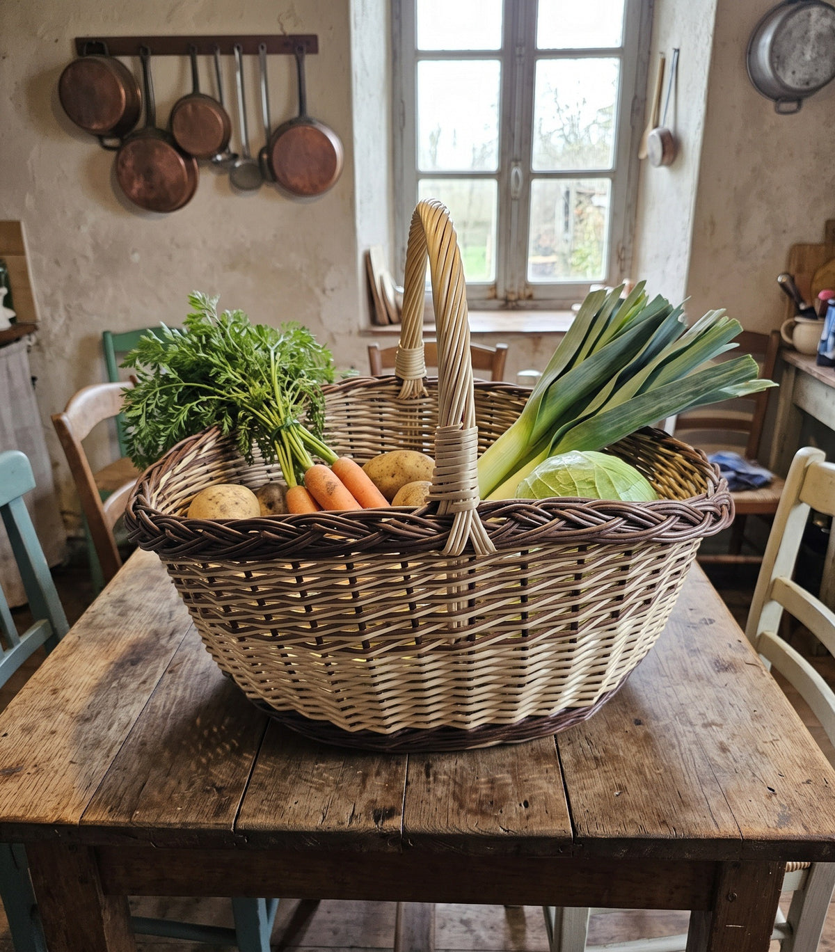 Large oval willow market basket with handle full of carrots, leeks, potatoes and cabbage on a farmhouse kitchen table