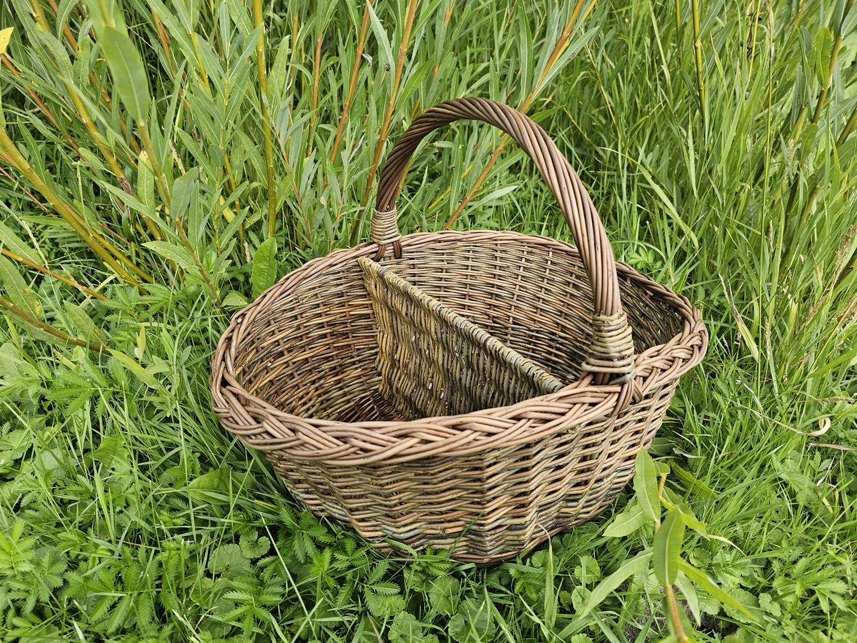 Handwoven willow picnic basket with an arched handle and a woven centre divider forming two compartments, set in tall grass.