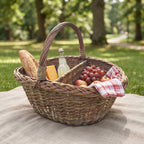Two compartment willow picnic basket packed with a baguette, cheese, grapes, lemonade and a checked napkin on a linen blanket in a park.