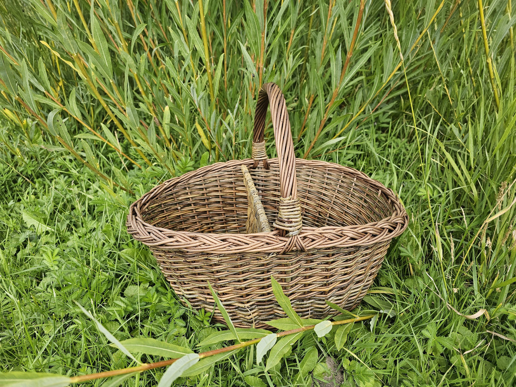 Oval willow market basket with arched handle and centre divider standing in tall grass beside willow shoots.