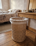 Round wicker waste basket with a bin liner, handwoven from willow, standing on a wooden floor beside a dressing table in a bedroom.