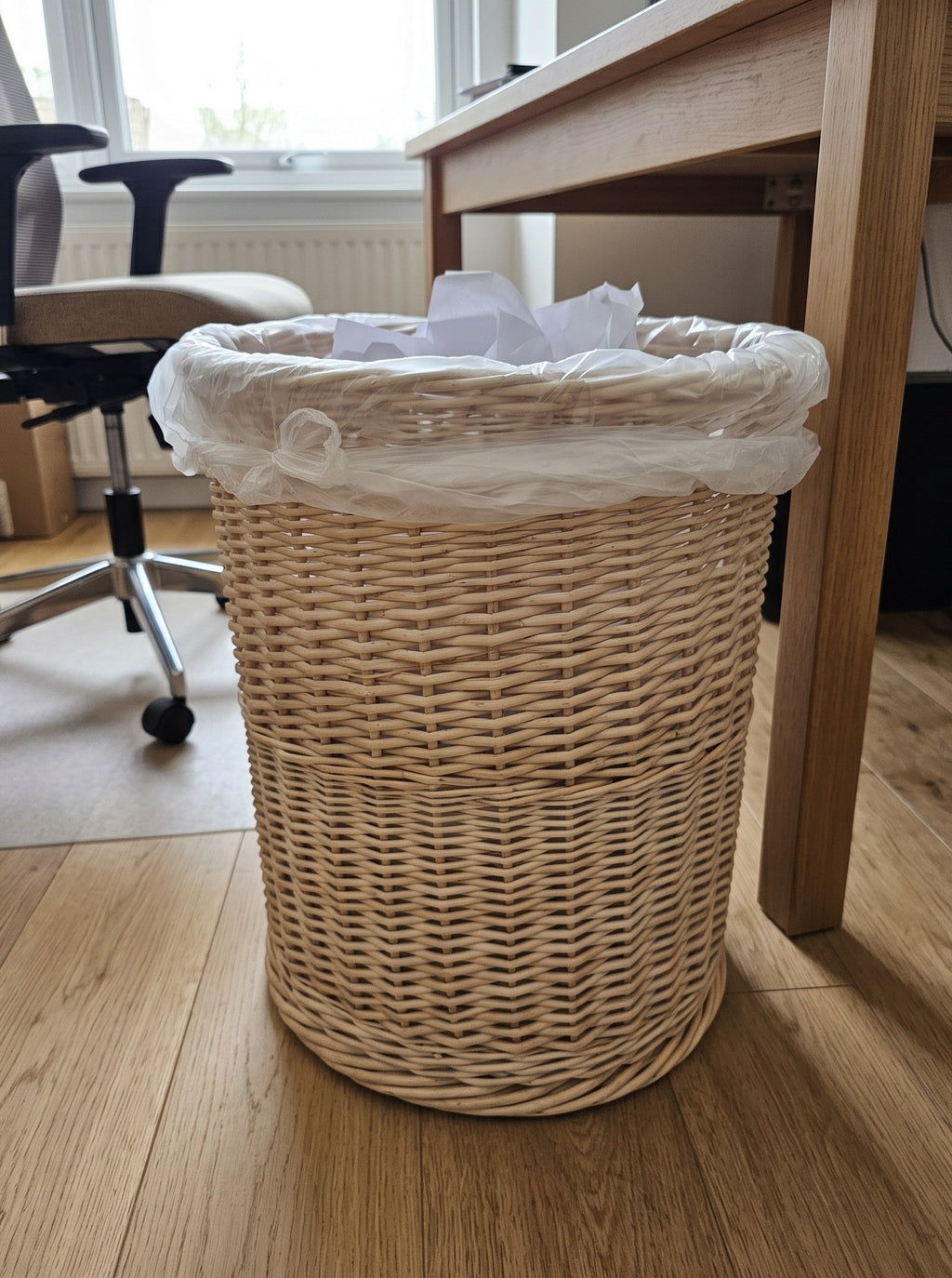 Handwoven willow trash can with paper inside used as an office waste bin under a wooden desk next to an office chair.