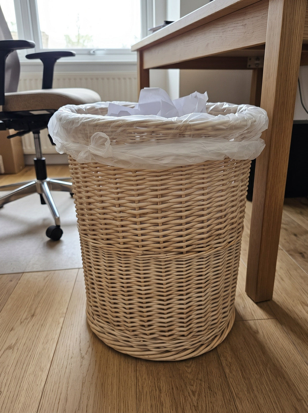 Handwoven willow trash can with paper inside used as an office waste bin under a wooden desk next to an office chair.