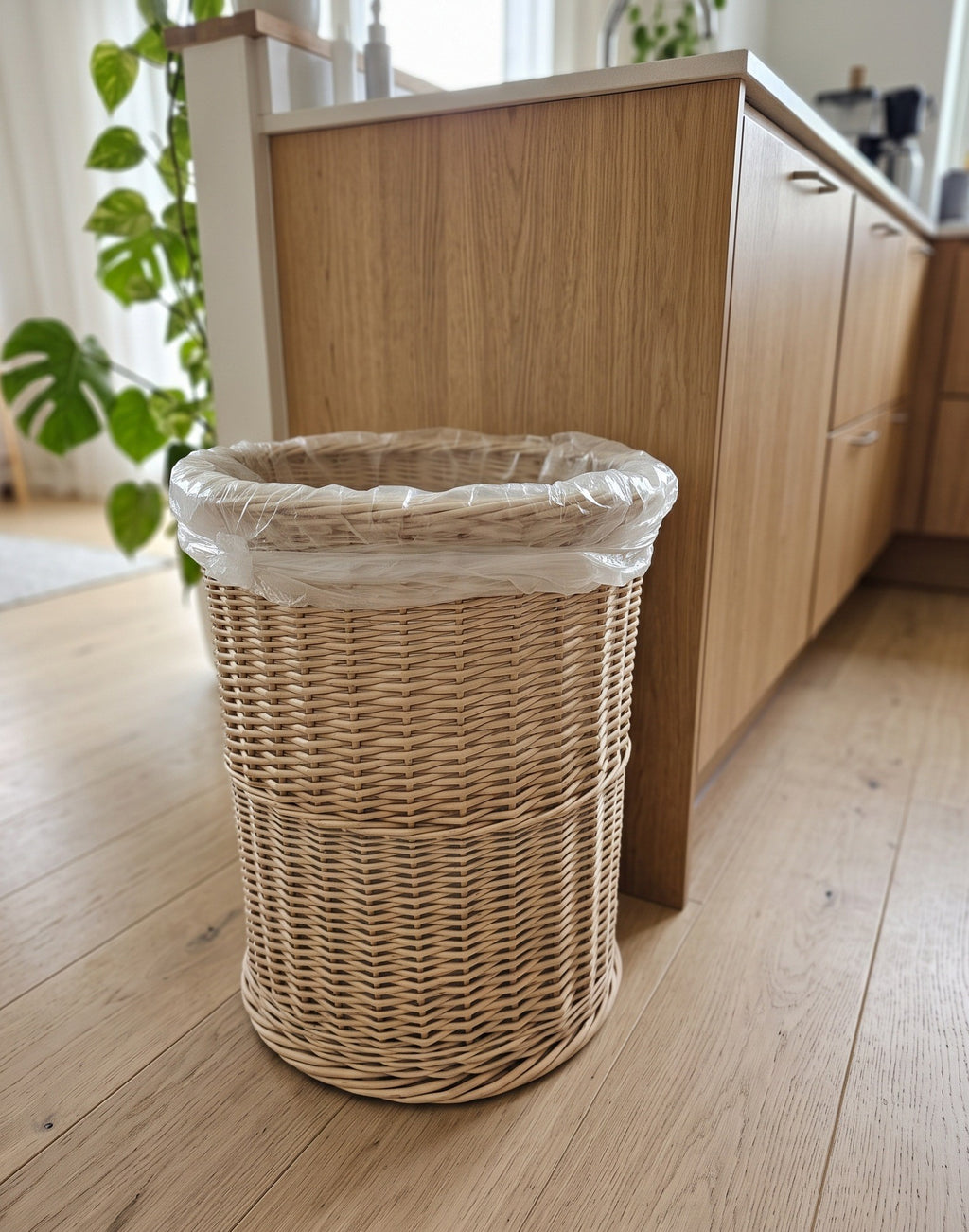 Round wicker kitchen bin with a liner standing on the floor beside wooden kitchen cabinets and a houseplant.