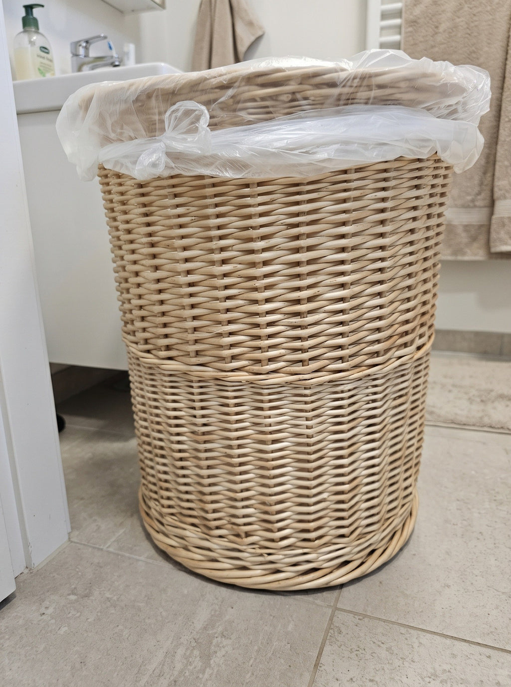 Round handwoven willow bathroom trash can with a bin bag inside on a stone tile floor next to a washbasin and towels.