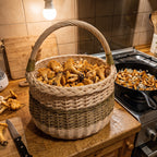 Willow mushroom basket filled with chanterelles on a wooden kitchen counter next to a pan of mushrooms frying on the stove