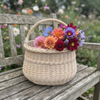 Medium willow basket with a braided rim and arched handle holding a bouquet of dahlias and asters on a weathered garden bench