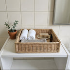 Shallow wicker storage tray with a wooden base holding rolled towels, a bar of soap and a bottle on a white bathroom shelf
