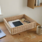 Rectangular willow desk organizer tray holding a notebook, papers, glasses and a tape roll on a wooden desk by a window