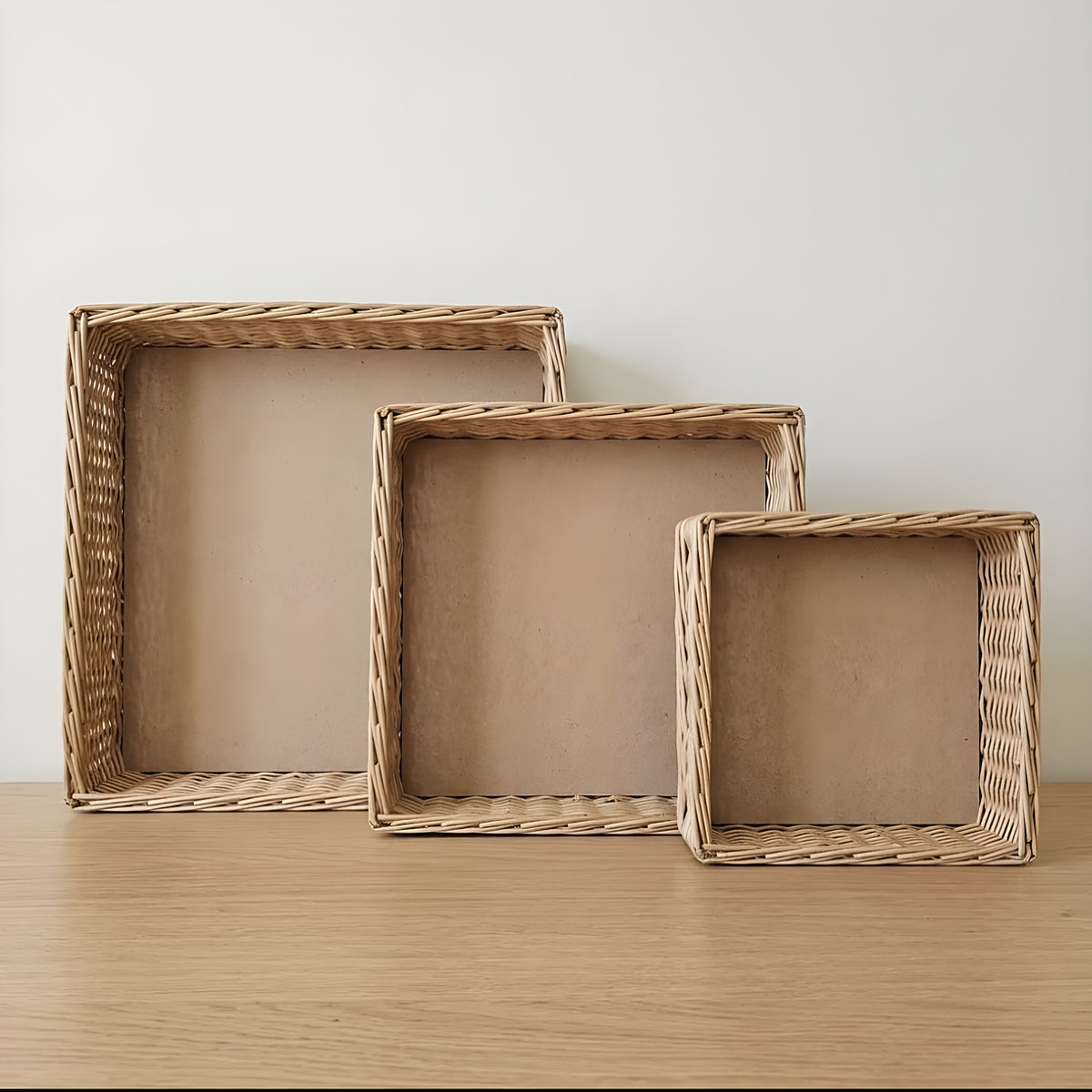 Three shallow handwoven willow storage trays in different custom sizes standing upright on a wooden surface against a white wall
