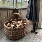 Large mushroom foraging basket handwoven from willow, filled with freshly picked porcini on a wooden porch floor beside rubber boots and a raincoat.