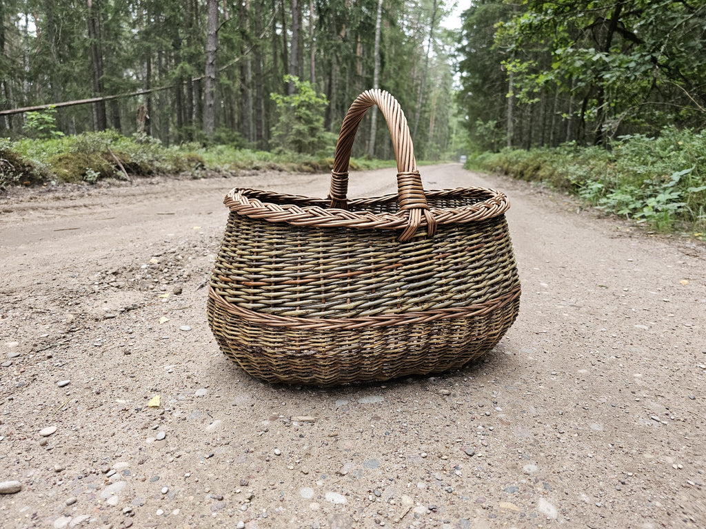 Willow foraging basket with an oval base, central arched handle and a braided rim, standing in the middle of a sandy forest road.