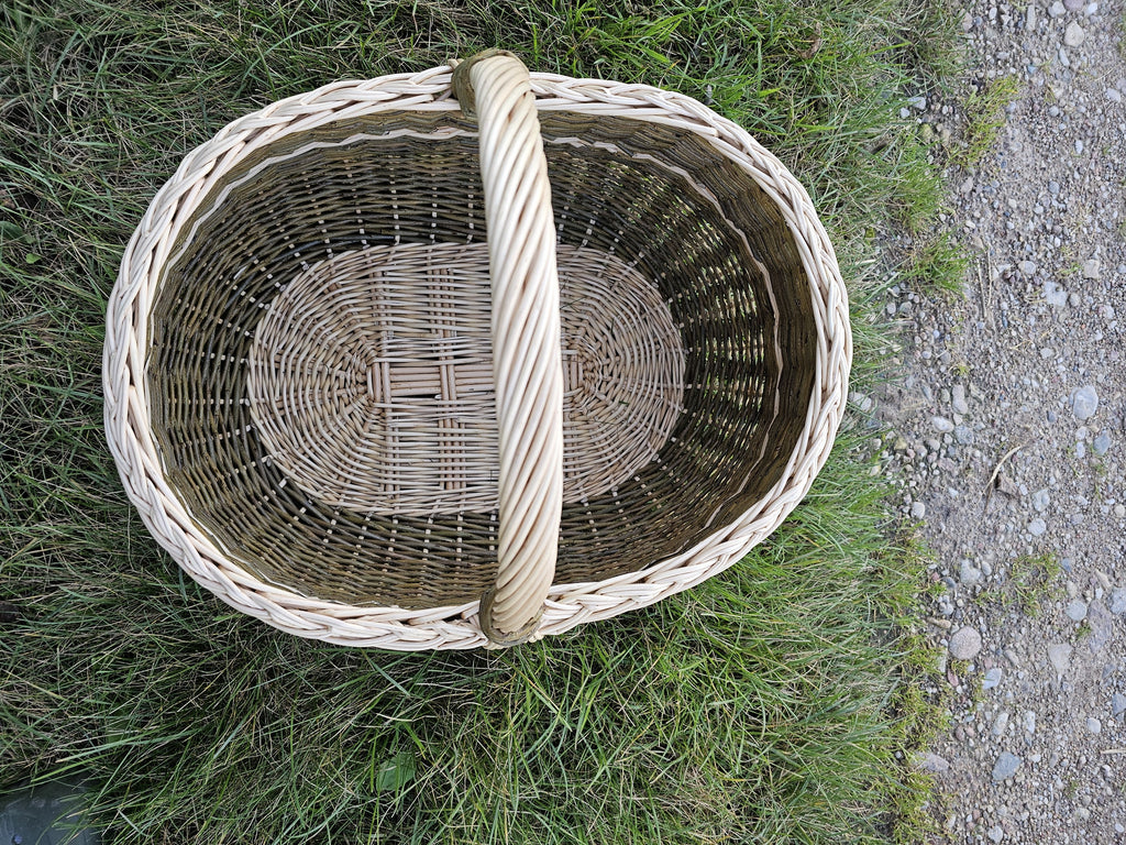 Top view into the large oval willow market basket on the grass, showing the woven oval base, braided rim and handle.