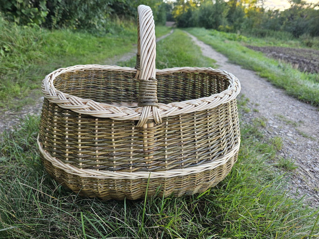 Deep oval foraging basket with a braided rim and contrasting woven bands standing on grass beside a country gravel road.