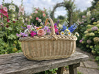 Flacher Erntekorb mit gedrehtem Henkel voller Rosen, Margeriten und Rittersporn auf einer Gartenbank zwischen Blumenbeeten