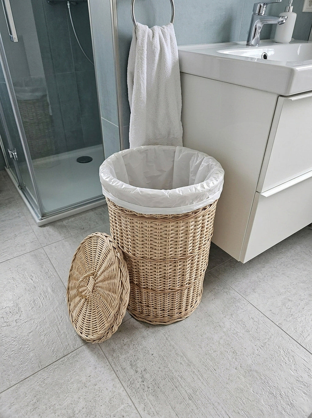Round wicker waste basket with lid used as a bathroom bin with a liner bag, lid leaning against it beside a shower and vanity