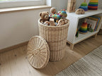 Round handwoven willow basket with lid used as toy storage, filled with a teddy bear and wooden toys in a child's room