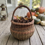 Round handwoven wicker basket with a braided rim holding pine cones and autumn leaves on a wooden porch beside pumpkins and a lantern