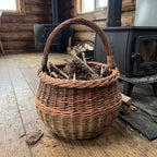 Round willow gathering basket with an arched handle filled with kindling and pine cones beside a cast iron stove in a log cabin