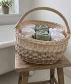 Oval willow storage basket with handle holding rolled bath towels on a wooden stool beside a bathtub, used as linen storage.