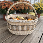 Oval wicker harvest basket holding mini pumpkins, gourds and pine cones on a porch with potted autumn chrysanthemums behind.