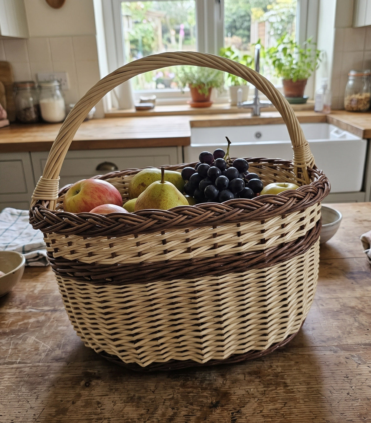 Handwoven willow basket with handle filled with apples, pears and grapes on a farmhouse kitchen counter by the sink and window.
