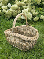 Oval wicker basket with a braided rim and arched handle sitting on the grass in front of flowering hydrangea bushes
