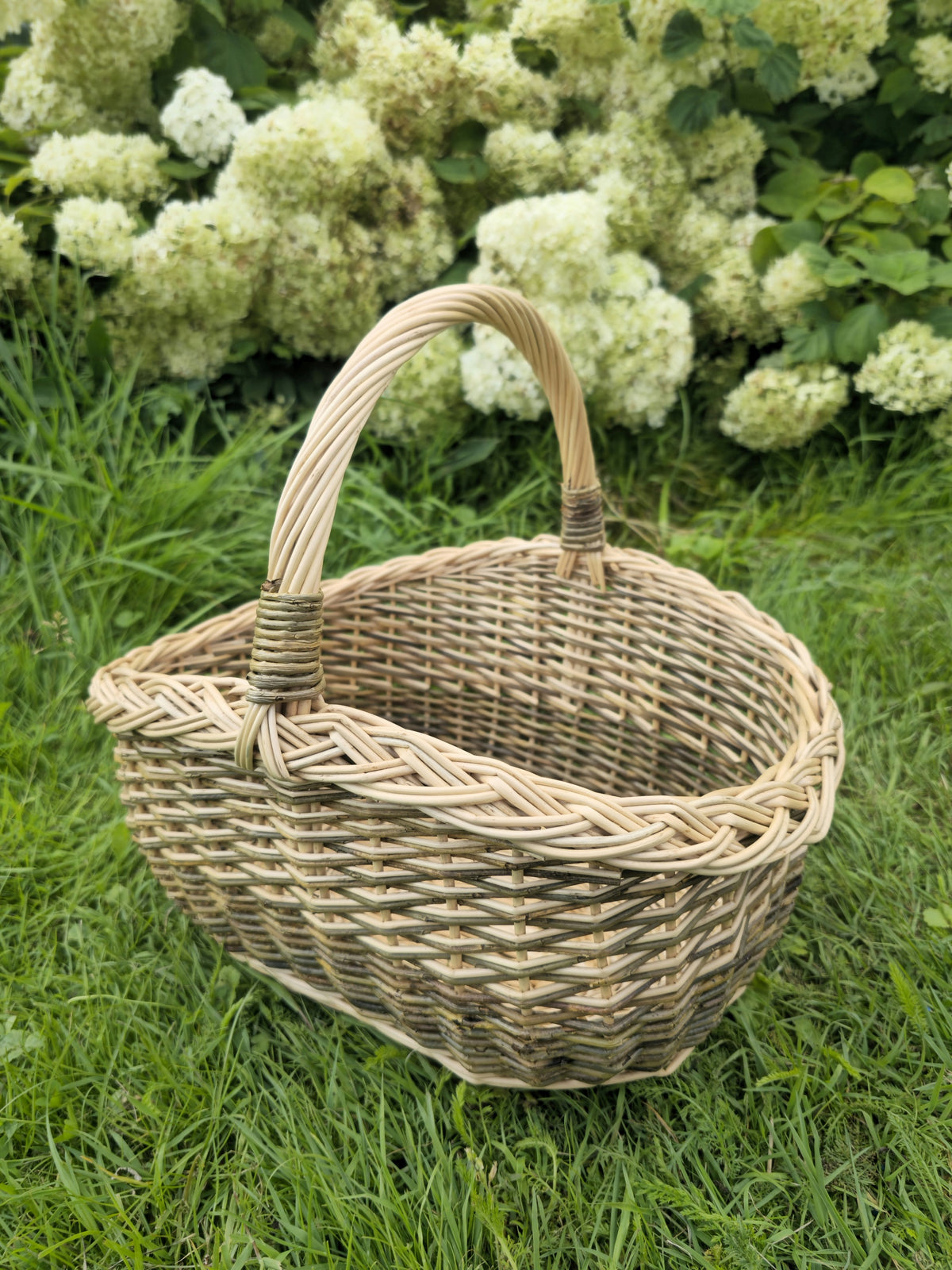 Oval wicker basket with a braided rim and arched handle sitting on the grass in front of flowering hydrangea bushes