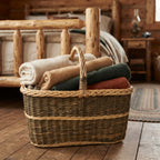 Large blanket basket with handle filled with folded wool throws, handwoven from willow, on the wooden floor of a log cabin bedroom.