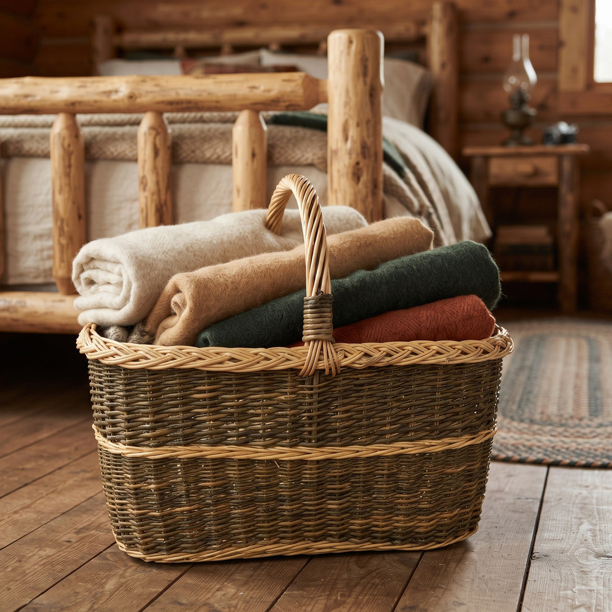 Large blanket basket with handle filled with folded wool throws, handwoven from willow, on the wooden floor of a log cabin bedroom.