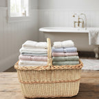 Rectangular wicker bathroom basket with handle holding stacked folded towels on a wooden bench beside a vintage bathtub.