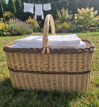 Large rectangular wicker laundry basket with handle filled with folded white sheets on the lawn with laundry drying behind.