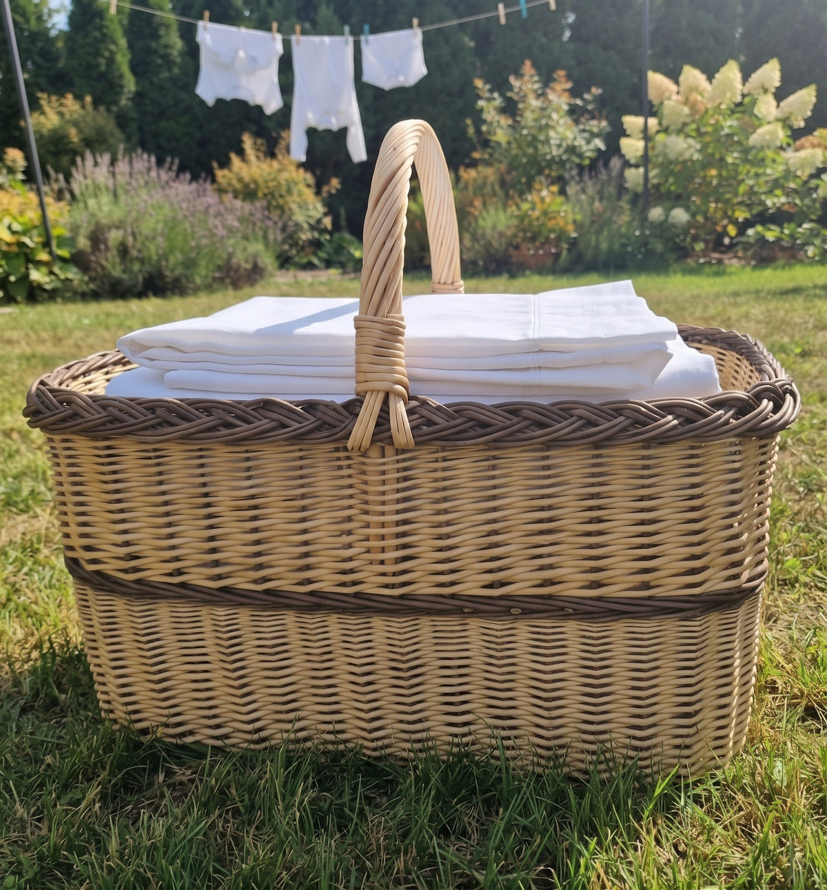 Large rectangular wicker laundry basket with handle filled with folded white sheets on the lawn with laundry drying behind.