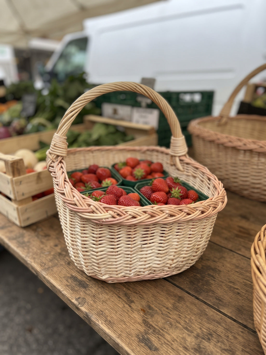 Rectangular wicker market basket holding punnets of fresh strawberries on a wooden farmers market stall with produce crates behind.