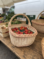 Rectangular wicker market basket holding punnets of fresh strawberries on a wooden farmers market stall with produce crates behind.