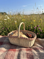 Willow picnic basket with handle holding bread and grapes on a checked wool blanket in a summer meadow of wildflowers.
