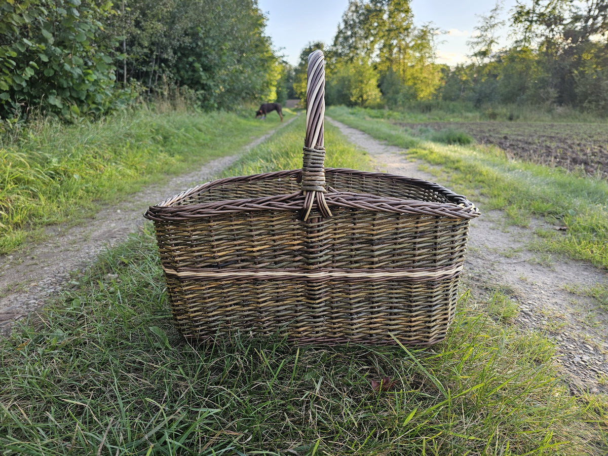 Großer Holzkorb mit hohem gedrehtem Henkel mitten auf einem grasbewachsenen Feldweg bei Sonnenuntergang