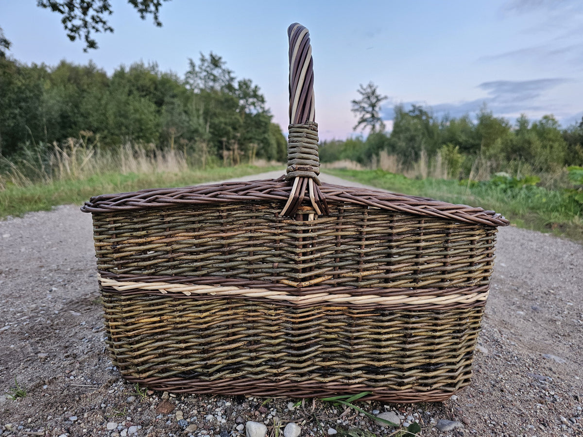 Seitenansicht des großen Holzkorbs mit hohem, aufrechtem Henkel auf einem Schotterweg unter dem Abendhimmel