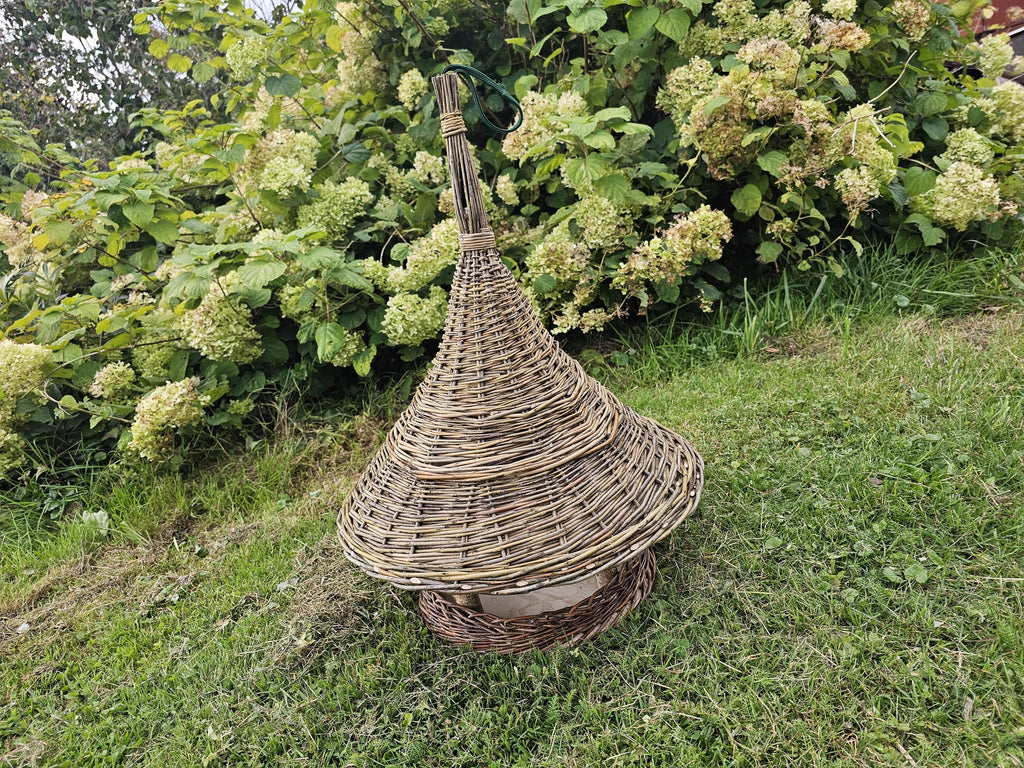 Handwoven willow bird table with a woven conical roof standing on a sloping lawn beside a large hydrangea bush