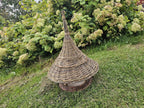 Handwoven willow bird table with a woven conical roof standing on a sloping lawn beside a large hydrangea bush